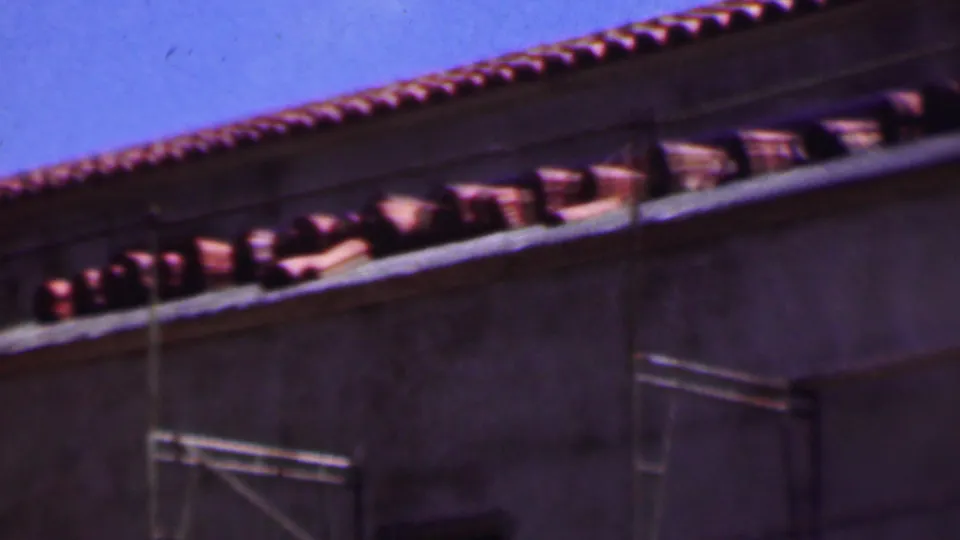 1956 Dallas 8mm frame with curved terracotta tiles along the roofline, a pale wall surface, and metal scaffolding crossing the building exterior.