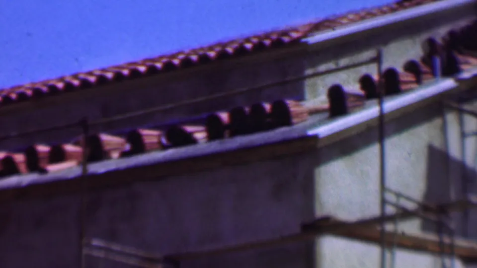 1956 Dallas 8mm frame showing a closer angle on terracotta roof tiles, dark roofline openings, a pale wall corner, and scaffold poles.