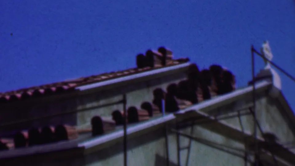1956 Dallas 8mm frame showing the upper building exterior with terracotta roof tiles, visible scaffolding, a pale wall, and open blue sky.