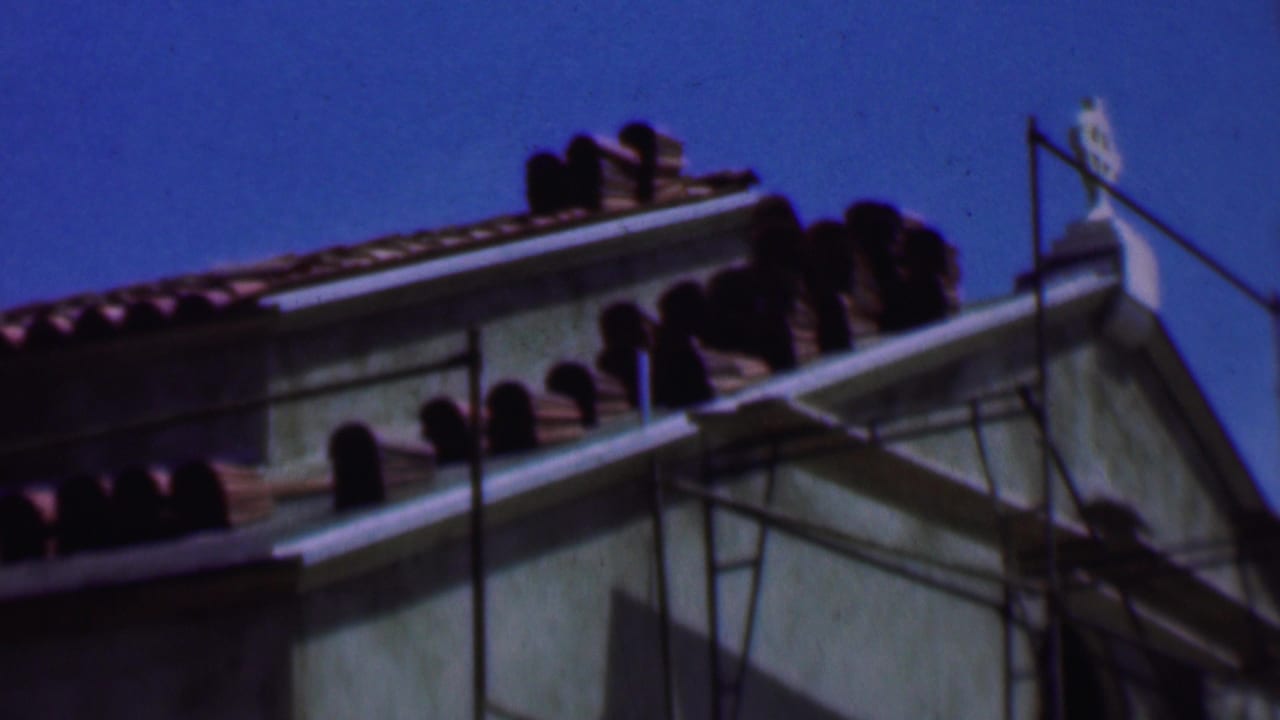 1956 Dallas 8mm frame showing a pale stucco building roofline with red-brown tile, scaffold poles, and a clear blue sky.