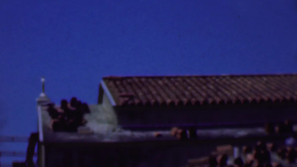 1956 Dallas 8mm frame showing a long red-brown tiled roof, pale stucco gable edge, blue sky, and dark construction elements below.