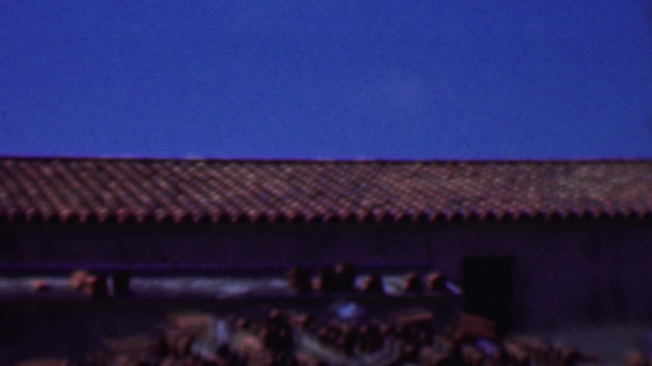 1956 Dallas 8mm frame showing a low building roofline with red clay tiles, dark wall openings, and a saturated blue sky.