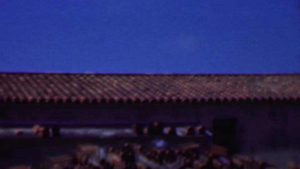 1956 Dallas 8mm frame showing a low building roofline with red clay tiles, dark wall openings, and a saturated blue sky.