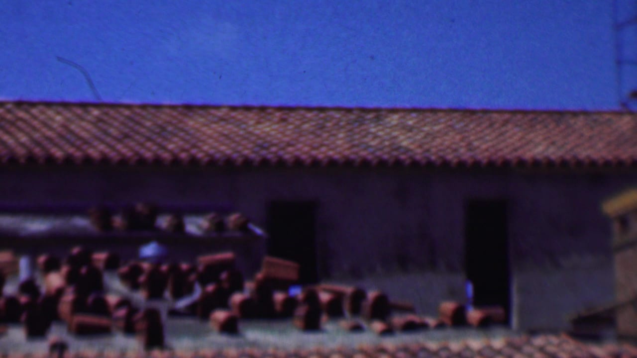1956 Dallas home-movie frame showing a red tile roof, dark doorways, and stacks of clay bricks or terracotta tiles near the building.
