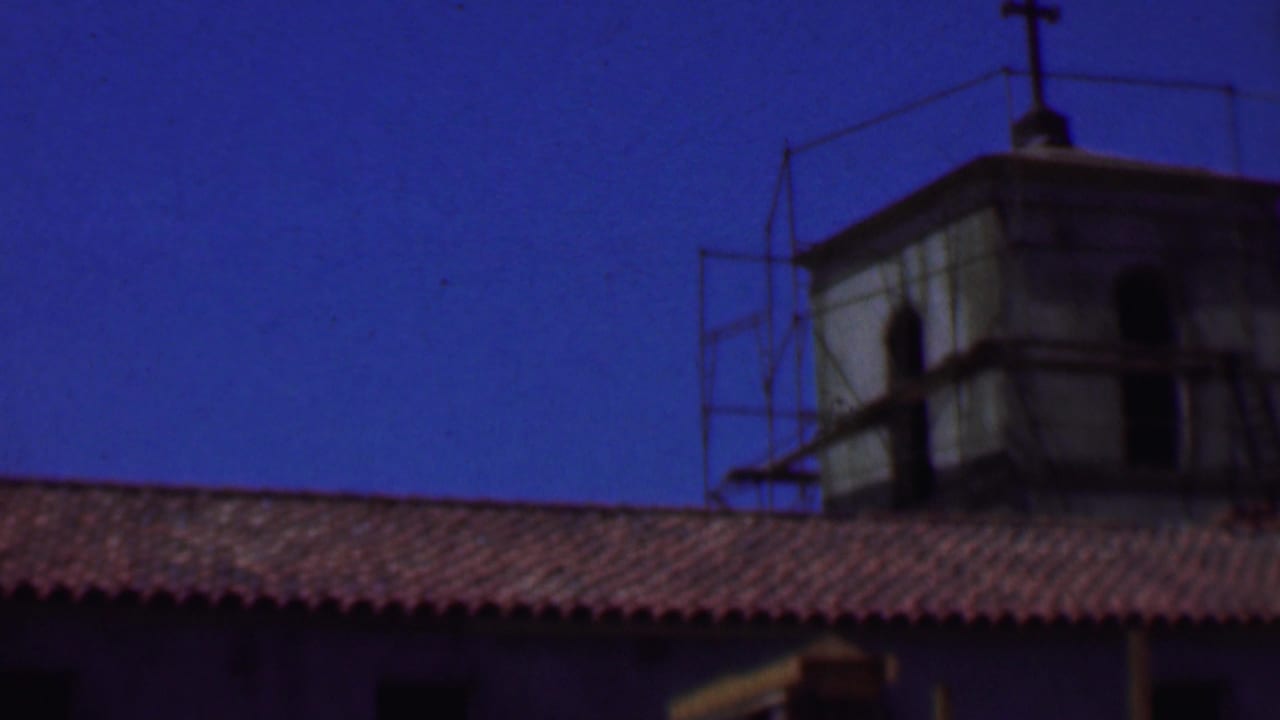 1956 Dallas home-movie frame showing an older stucco building with red roof tiles, tower structures, and metal scaffolding against a saturated blue sky.