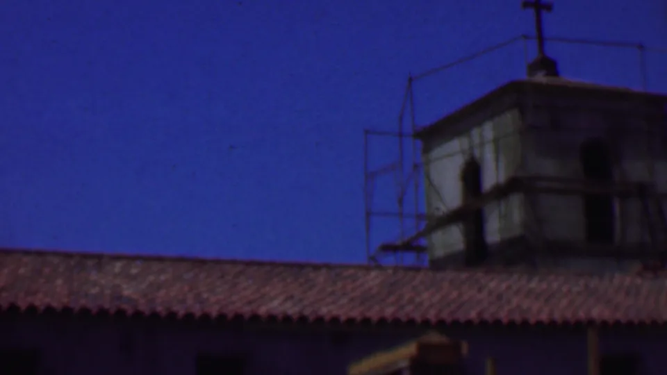1956 Dallas home-movie frame showing an older stucco building with red roof tiles, tower structures, and metal scaffolding against a saturated blue sky.