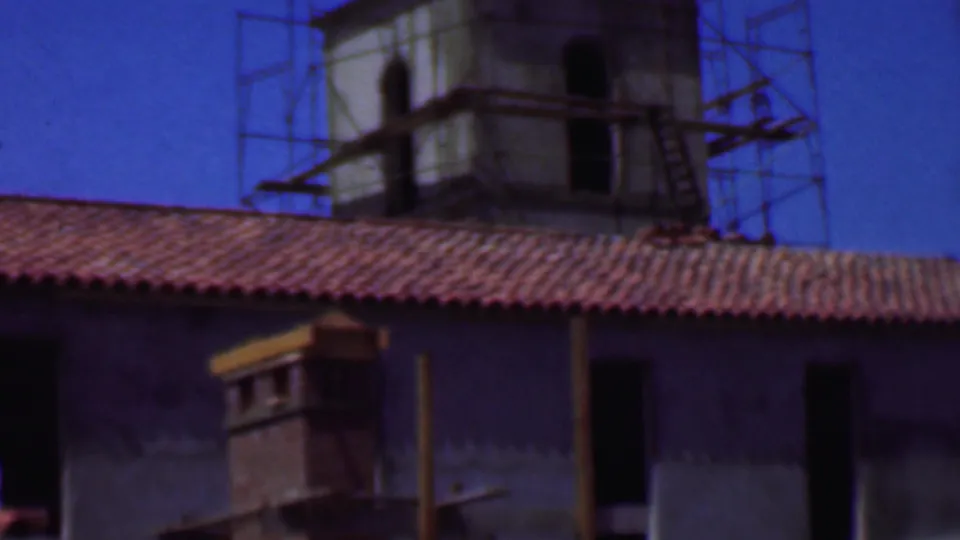 1956 Dallas home-movie frame showing a stucco facade below a red tile roof, with a scaffolded tower, dark door opening, and strong roofline shadow.