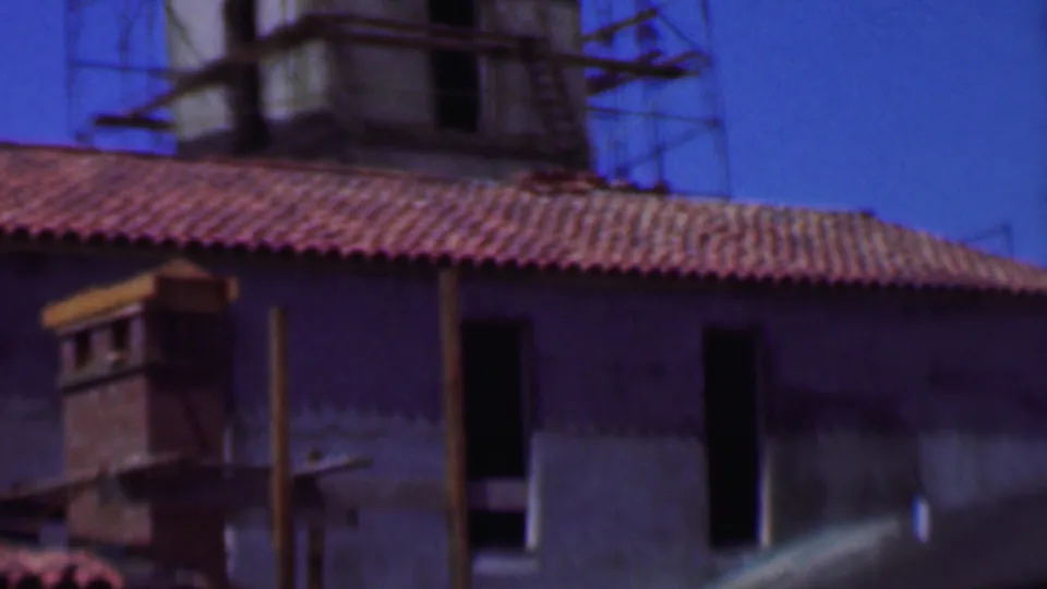 1956 Dallas frame showing the stucco building from a slightly shifted angle, with red tiles, tower scaffolding, dark window openings, and soft 8mm color.