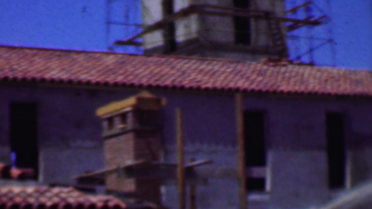1956 Dallas 8mm frame showing a red clay tile roof, unfinished masonry wall, dark window openings, and scaffolding around a tower-like structure in the background.