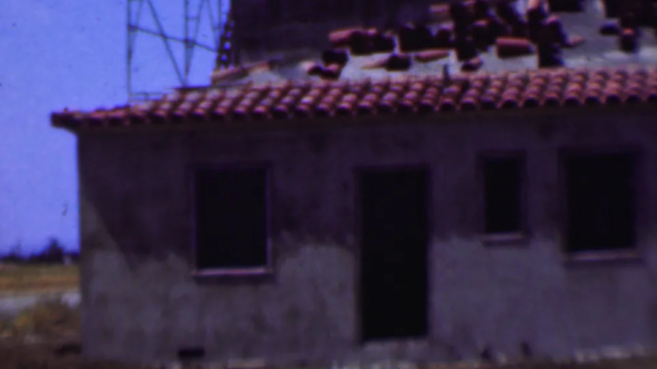 1956 Dallas frame of a partially finished building with a red tile roof, dark door and window openings, stacked roof tiles, and scaffold poles nearby.