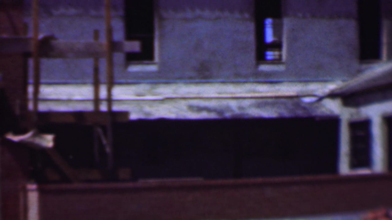 Dallas, Texas home-movie frame from the supplied 1956 clip showing an unfinished concrete or stucco building facade with dark window openings, scaffolding, and a low foreground barrier.