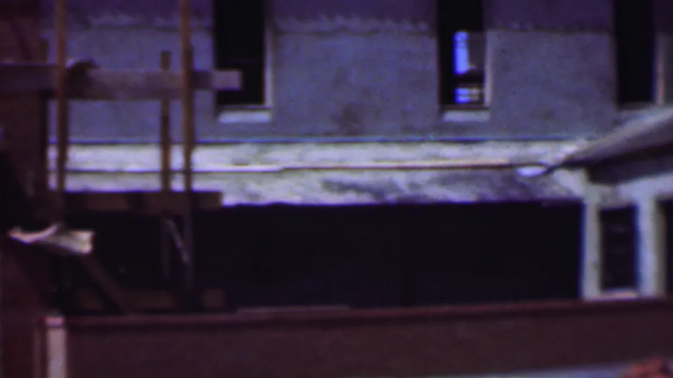 Dallas, Texas home-movie frame from the supplied 1956 clip showing an unfinished concrete or stucco building facade with dark window openings, scaffolding, and a low foreground barrier.