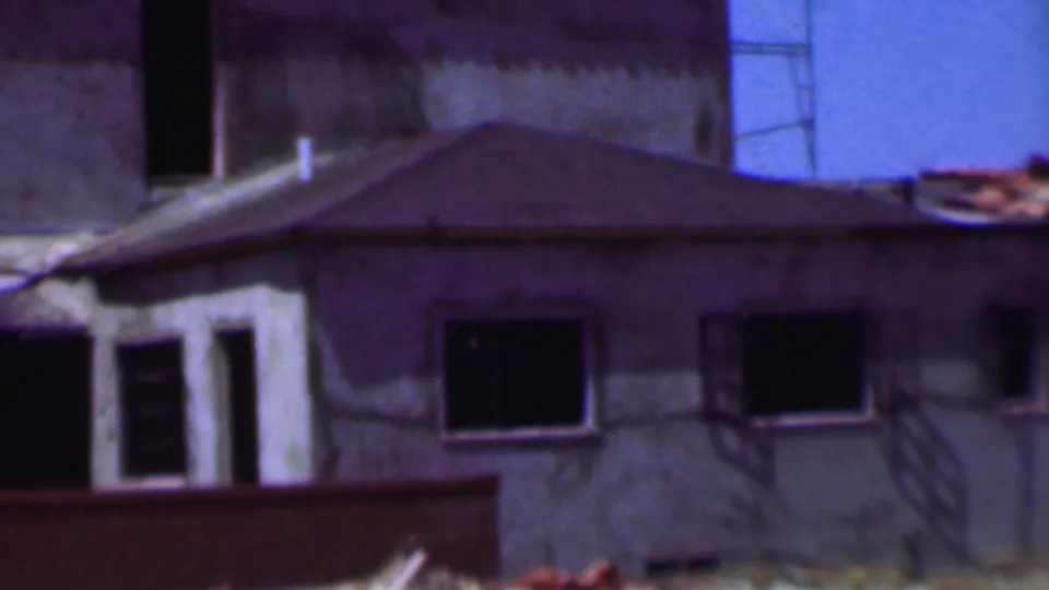 Frame from the 1956 Dallas home movie showing the side of an unfinished residential-scale structure with dark window openings, a low roof, and blue sky beyond.