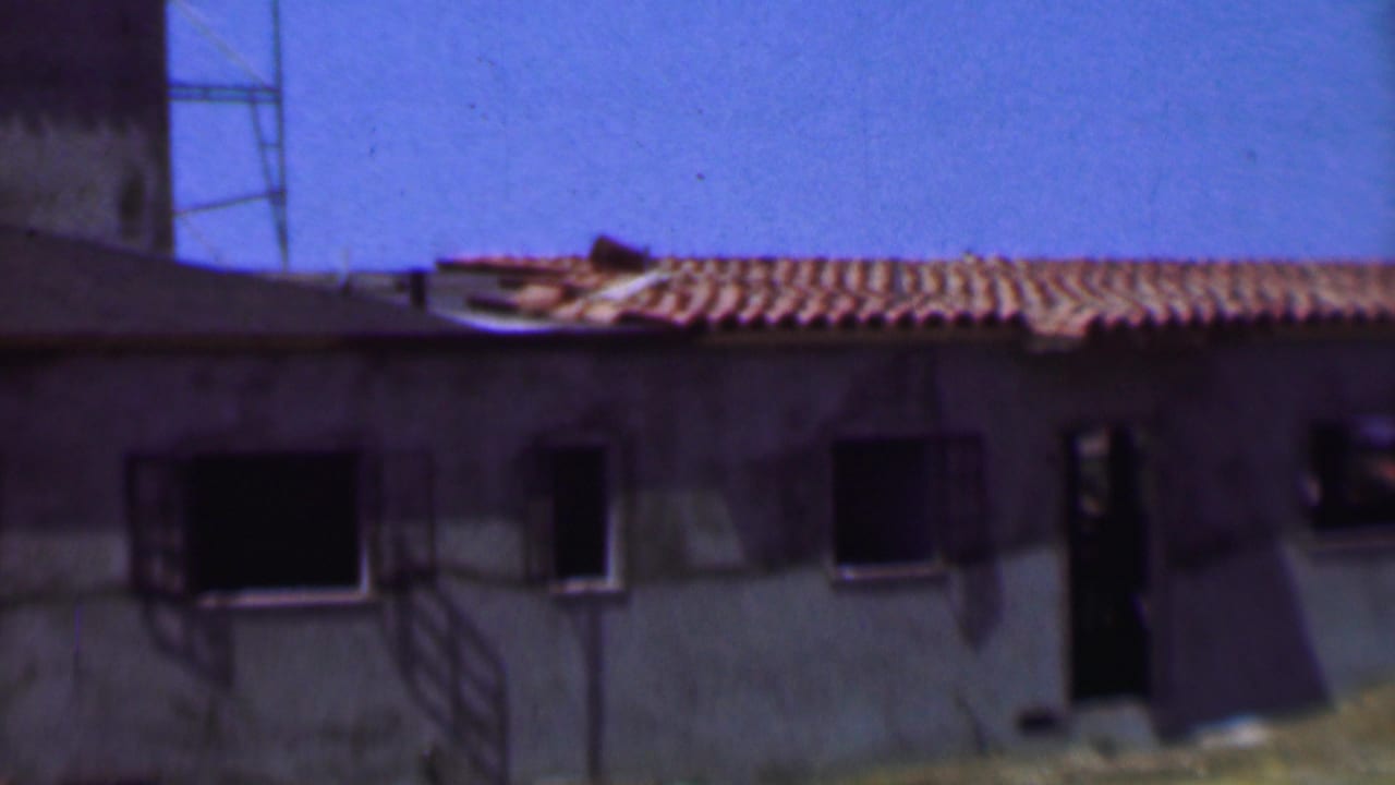 1956 Dallas home-movie frame showing a low unfinished concrete building with terracotta roof tiles, dark windows, and an open doorway beneath a clear blue sky.