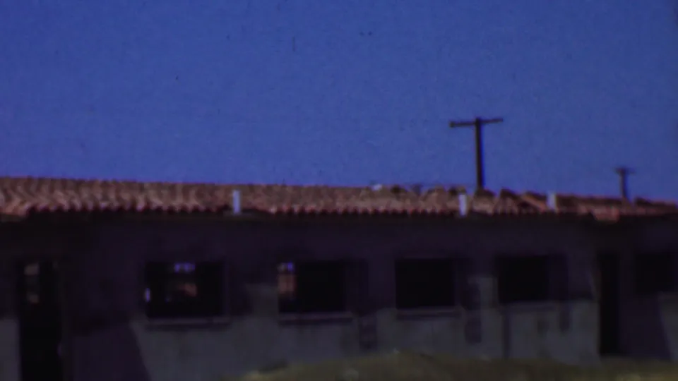 1956 Dallas home-movie frame showing the unfinished building roofline, dark window openings, bare ground, and a utility pole in the background.