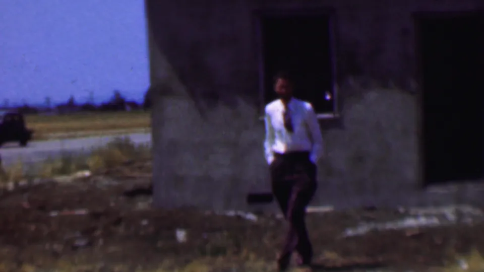 1956 Dallas frame showing a man in a white shirt and dark trousers walking past an unfinished concrete building, with a road and distant vehicle behind him.