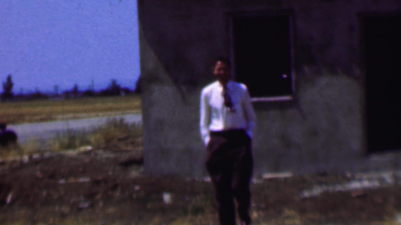In supported 1956 Dallas home-movie footage, a man in a white dress shirt and dark tie stands near a weathered concrete building with an open roadside landscape behind him.