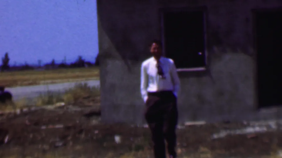 In supported 1956 Dallas home-movie footage, a man in a white dress shirt and dark tie stands near a weathered concrete building with an open roadside landscape behind him.