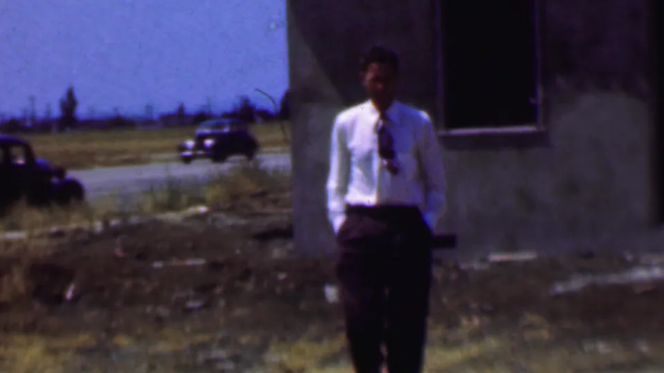 The man remains near the unfinished-looking building in 1956 Dallas footage, with older cars and a grassy roadside visible in the background.