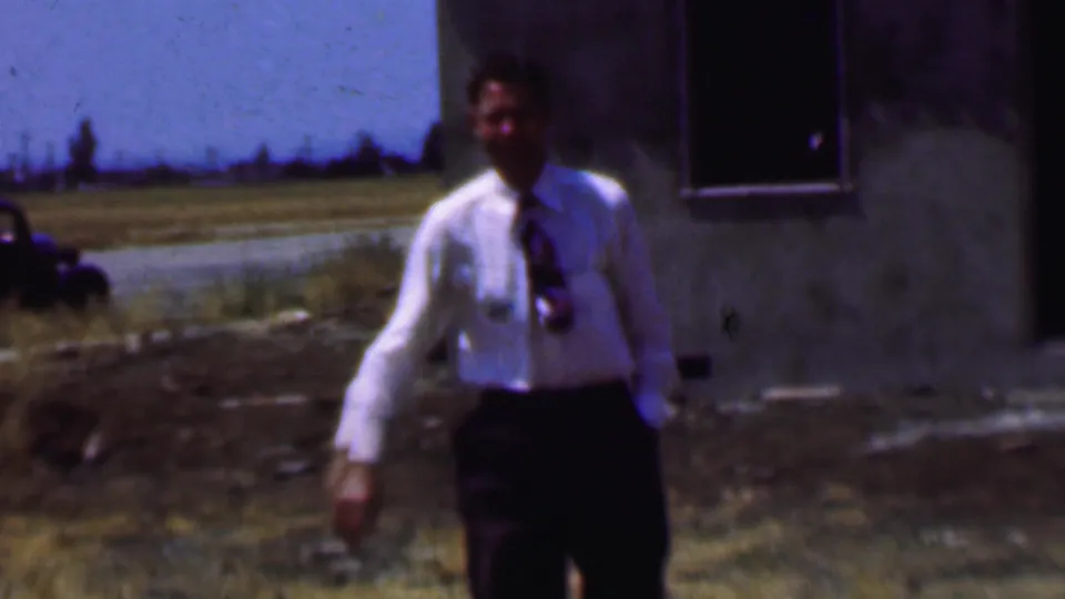 A man in a white dress shirt and tie walks toward the camera across a rough lot beside the concrete building, with roadside grass and an older car behind him.