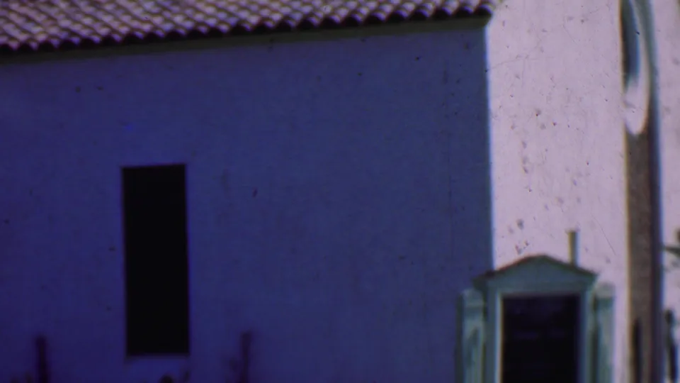 The final frame shows the pale exterior wall of the roadside building, including a dark vertical opening, roof tiles, and a small framed feature along the right edge.