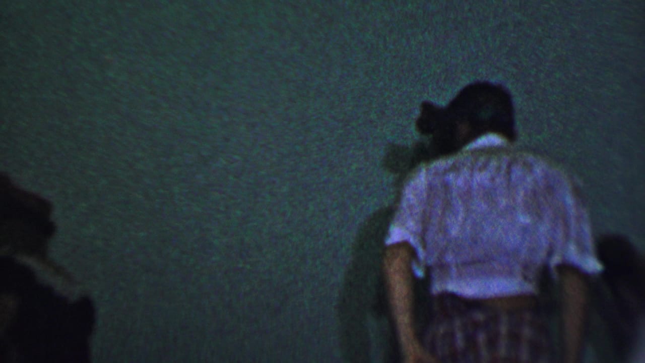 Guests at the Dallas party move through a dim interior space, with a woman in a light blouse and patterned skirt visible near the right side of the frame.