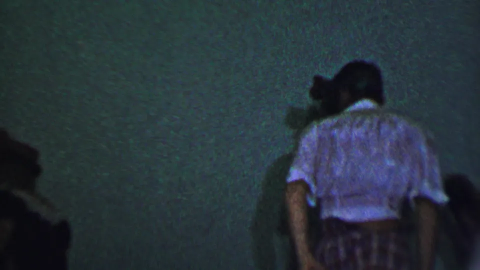 Guests at the Dallas party move through a dim interior space, with a woman in a light blouse and patterned skirt visible near the right side of the frame.