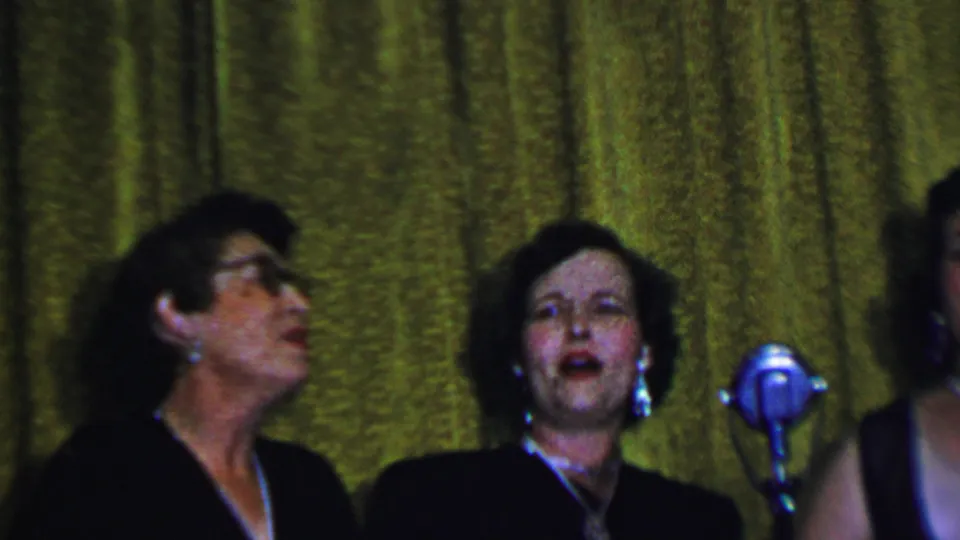 Two women sing or perform beside a vintage microphone against a textured gold curtain during the 1956 Dallas family party.