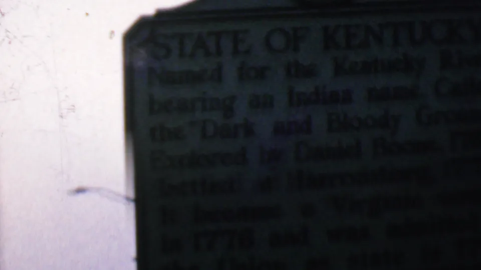 Handheld 8mm frame from Covington, Kentucky showing the Kentucky state marker shifted right, with inscription text partly legible and a blank sky area beside it.