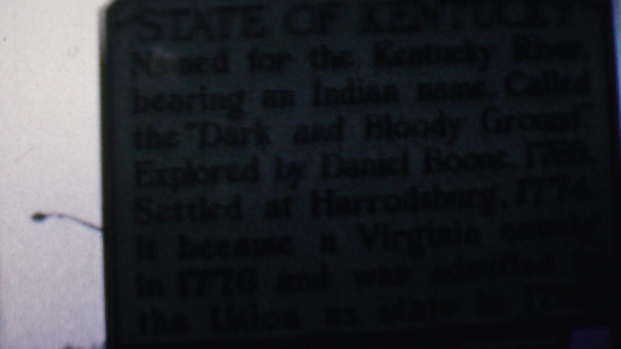Close 8mm frame of a dark green Kentucky historical marker in Covington, Kentucky, with the sign text mostly shadowed and a bright outdoor sky behind it.