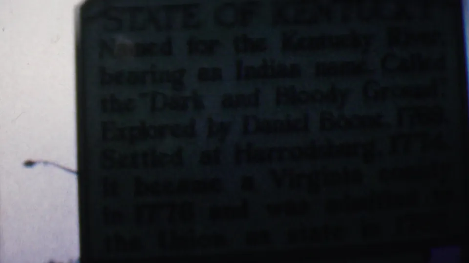 Close 8mm frame of a dark green Kentucky historical marker in Covington, Kentucky, with the sign text mostly shadowed and a bright outdoor sky behind it.