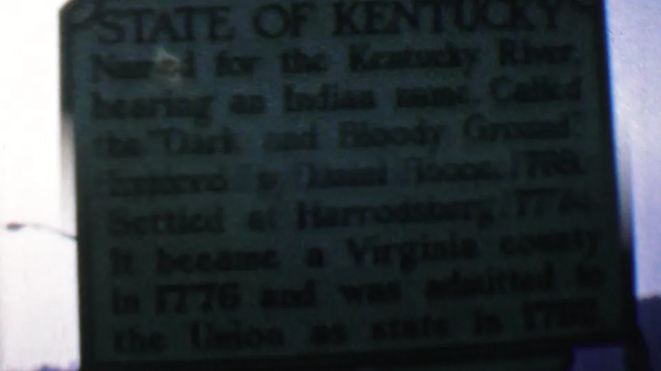 Handheld 8mm frame of the green Kentucky historical marker outdoors, with a street lamp at the left edge, pale sky behind the sign, and the marker text softened by exposure and focus.