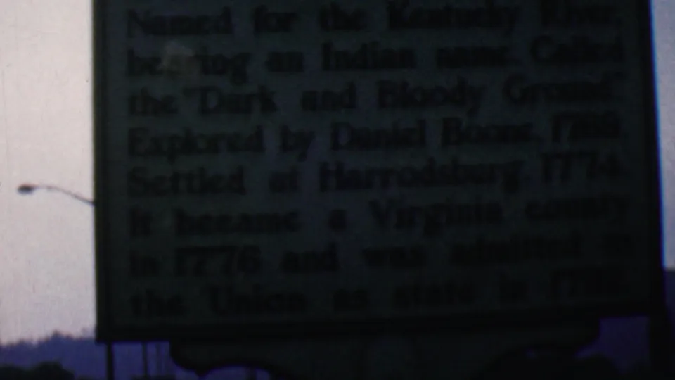 Close 1961 home-movie view of a dark green Kentucky historical marker in Covington, with dense white text, a visible street lamp, pale sky, and a low wooded ridge in the background.
