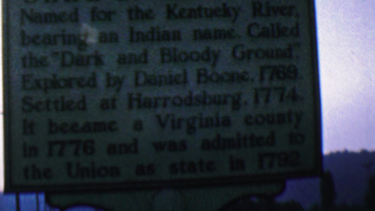 1961 Covington, Kentucky home-movie frame showing a dark historical marker about Kentucky history with a river barge passing behind it.