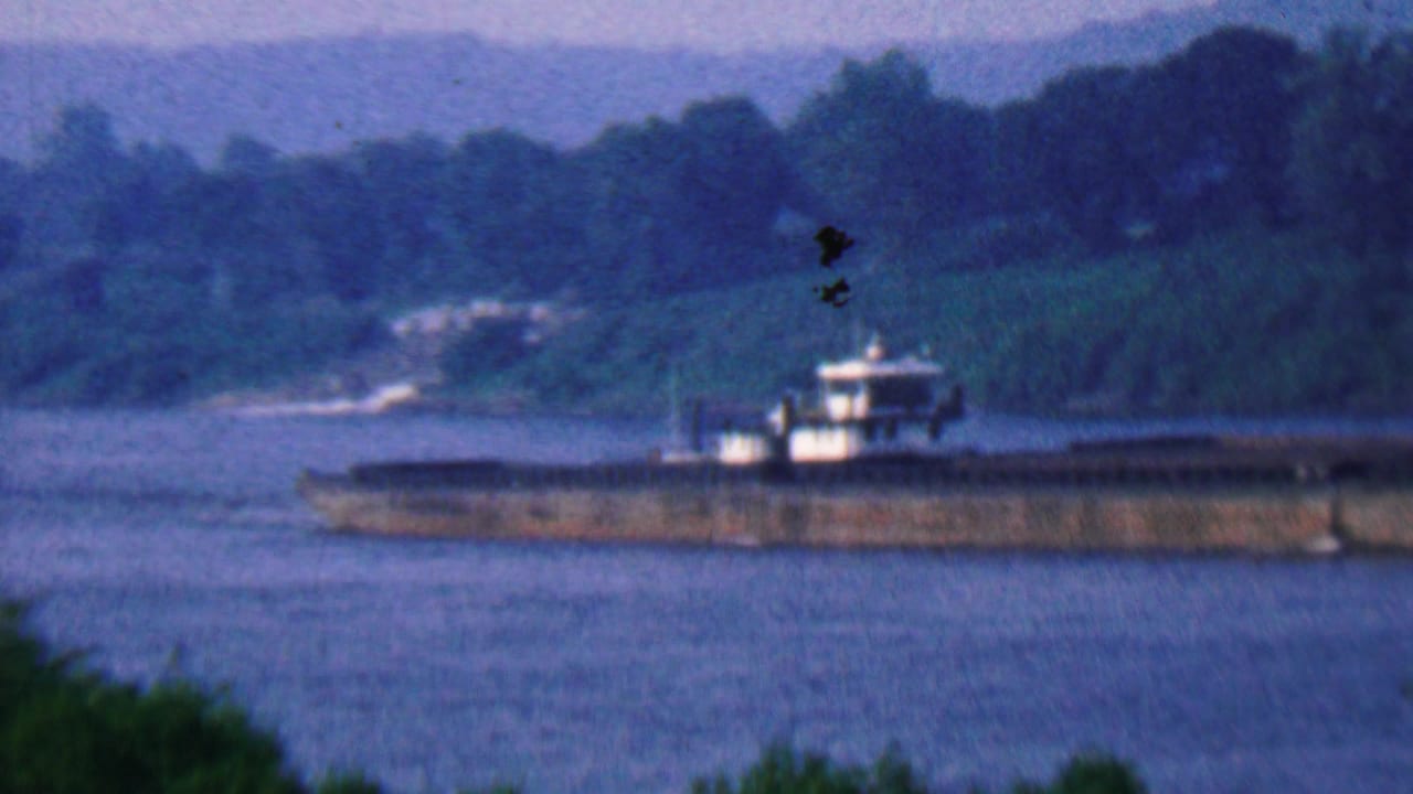 Long barge moving along a river with wooded hills behind it.