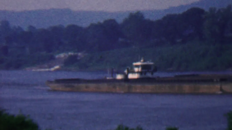 Soft 8mm frame of a barge and towboat on the river near Covington, Kentucky, with forested banks and hazy hills beyond.