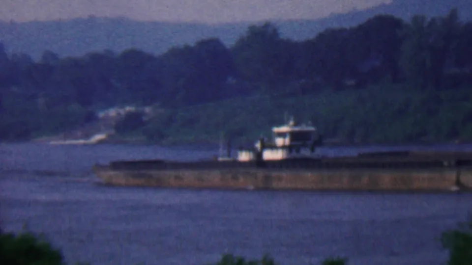 1961 home-movie frame showing a towboat and long barge on the Kentucky riverfront, with wooded slopes and a soft blue sky in the background.