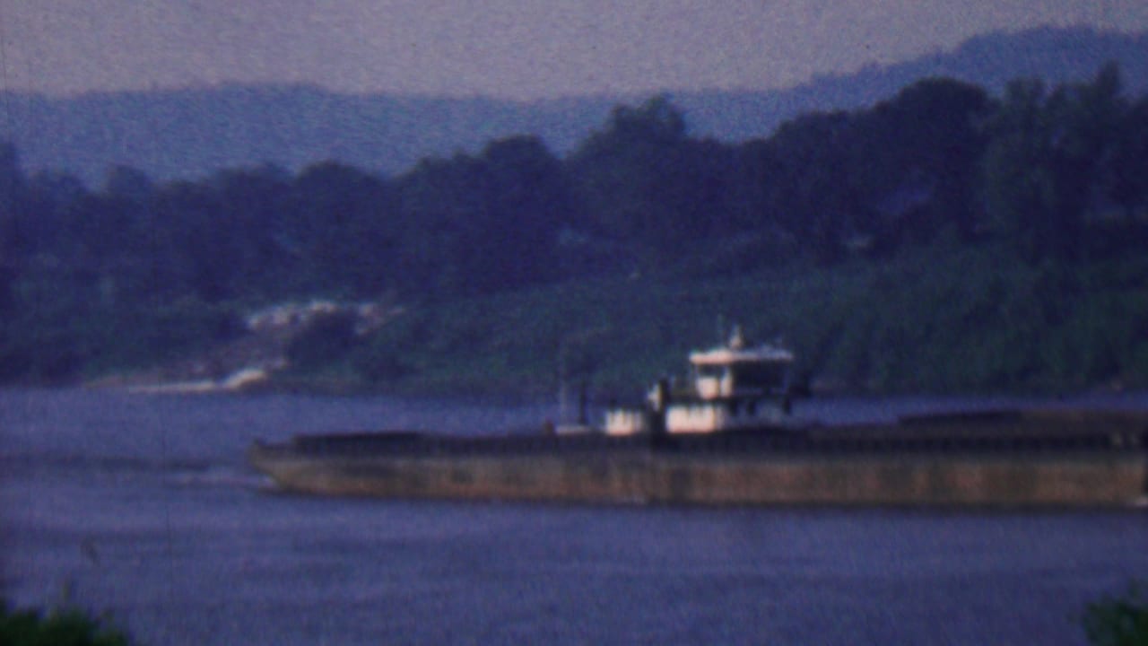 1961 Covington, Kentucky 8mm frame showing a long barge moving across a wide river at dusk, with dark wooded banks and distant hills behind it.
