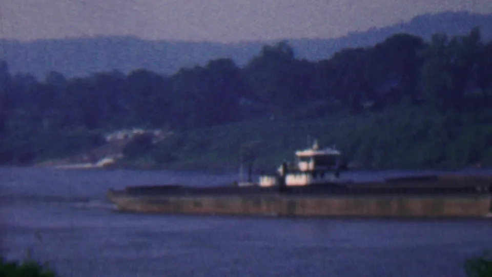1961 Covington, Kentucky 8mm frame showing a long barge moving across a wide river at dusk, with dark wooded banks and distant hills behind it.