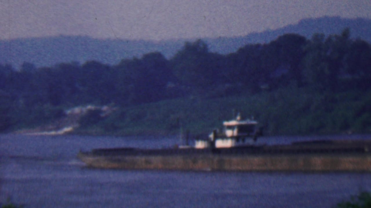 Barge continuing along the river with a tree-lined bank and hills beyond.