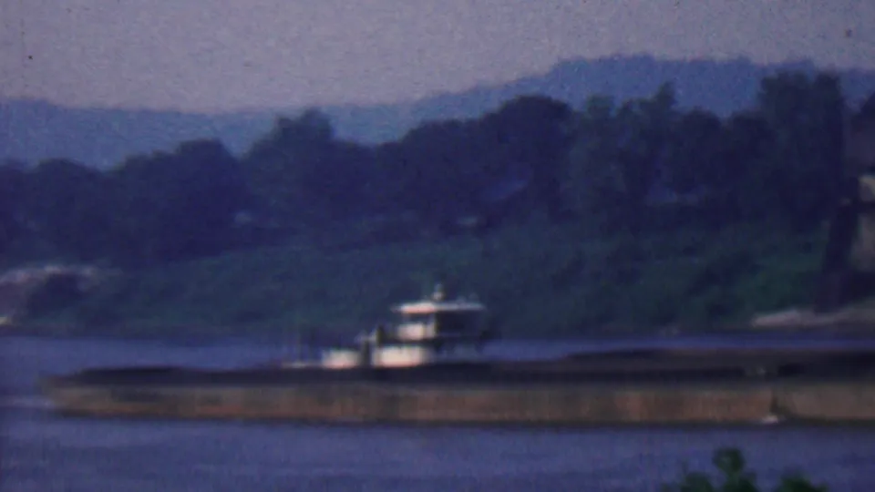 1961 Covington, Kentucky 8mm frame with the barge partly low in the composition, set against a dusky river, wooded shoreline, and hazy hills.
