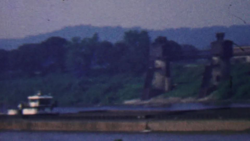 1961 Covington, Kentucky home-movie frame showing the barge near shore at dusk, with wooded banks, hills, and a bridge or industrial structure at the right side.