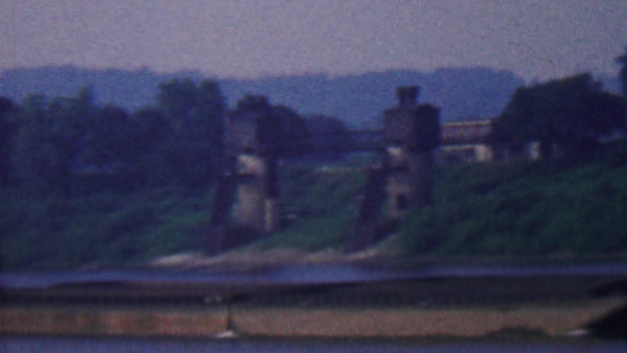 1961 8mm frame from metadata-identified Covington, Kentucky, showing a low barge on a river below green banks and distant bridge supports.