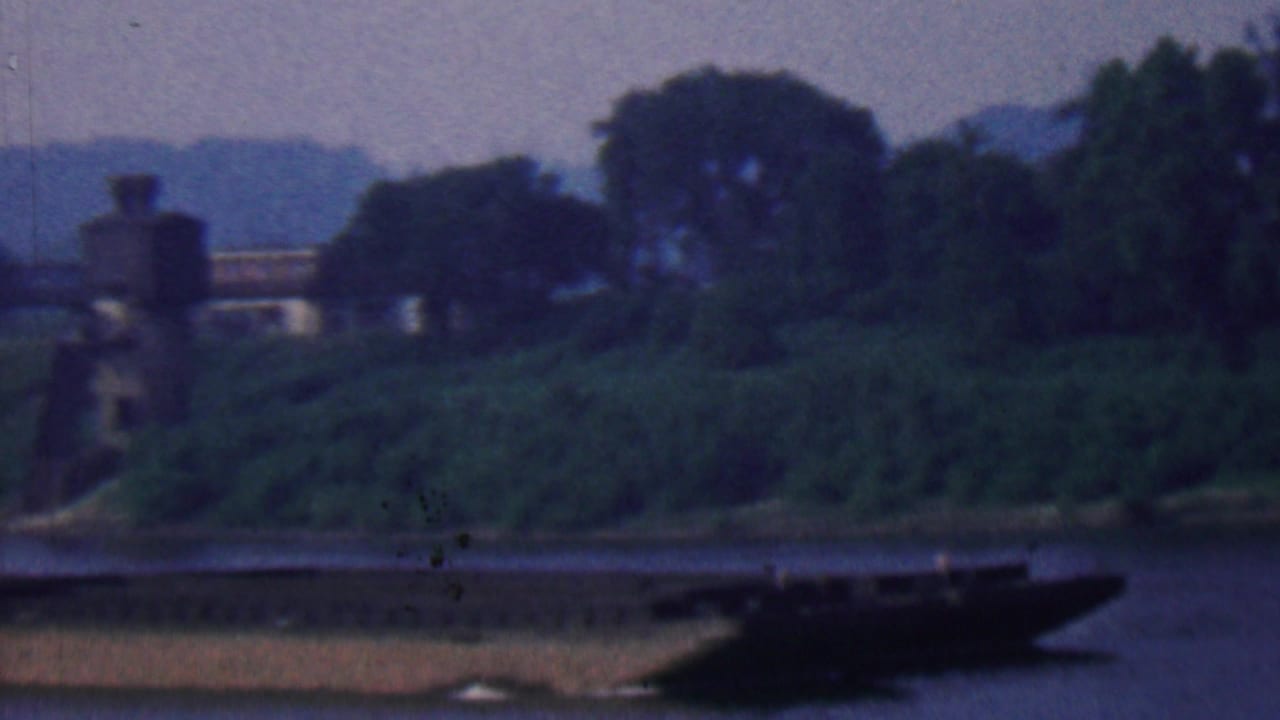 Green riverbank with distant bridge structure and a barge at the right edge.