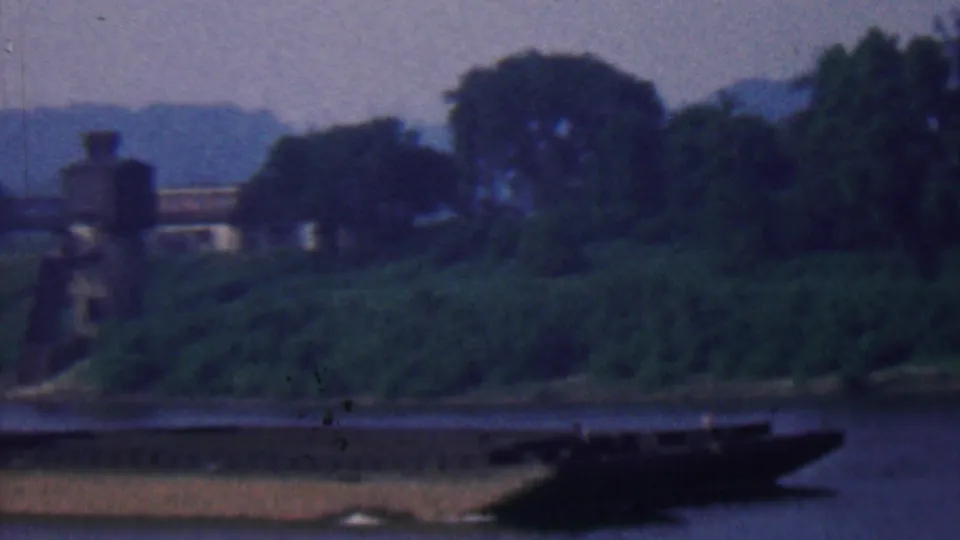 Soft 8mm view of a tree-lined riverbank and distant bridge structure, with a dark barge entering at the right edge in the Covington-area clip.
