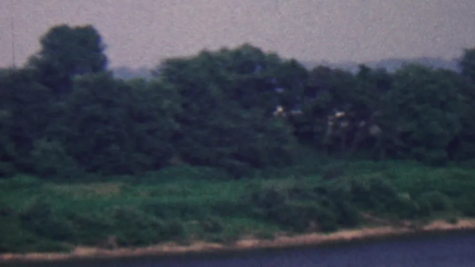 Covington, Kentucky 1961 8mm frame of a wooded shoreline, dark foreground water, distant hazy hills, and muted overcast sky with no visible people.