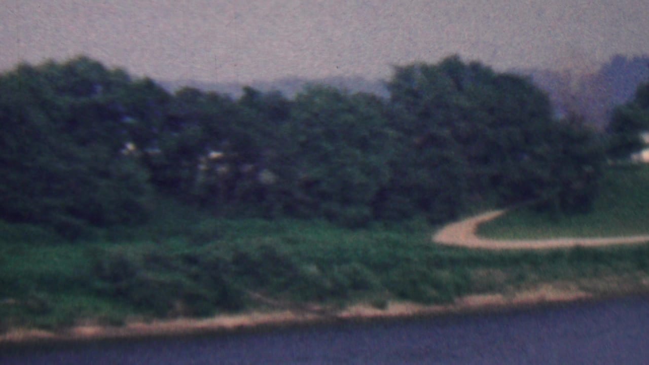 1961 Covington, Kentucky home-movie frame showing a soft, distant view across water toward a tree-lined bank with a pale winding path.