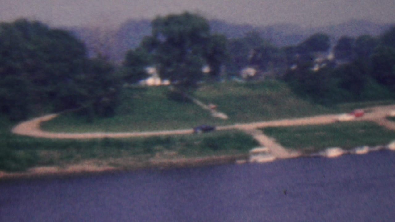 Continuation of the riverside view with paths, trees, cars, and shoreline visible.