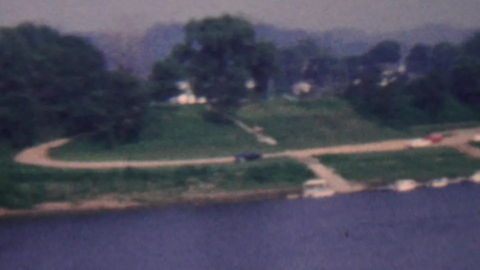 1961 Covington, Kentucky frame with a river in the foreground, shoreline paths, clustered trees, small cars, and distant houses on a hill.