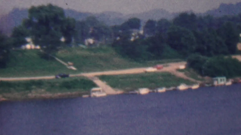 1961 Covington, Kentucky home-movie frame showing a broader shoreline with water below, a pale road, small cars, trees, and houses in the distance.